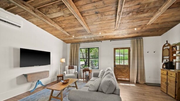Un salon avec un plafond en bois, un canapé gris, un téléviseur fixé au mur et une fenêtre avec des rideaux.