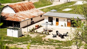 Il s’agit d’une propriété rurale comprenant une grande grange, un petit bâtiment blanc doté d’un toit en métal, ainsi qu’un espace patio aménagé avec des sièges d’extérieur.