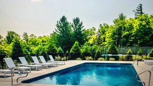 A rectangular pool with clear blue water, surrounded by a paved deck. There are lounge chairs and a fence with trees in the background.