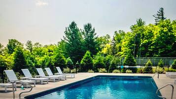 A rectangular pool with clear blue water, surrounded by a paved deck. There are lounge chairs and a fence with trees in the background.