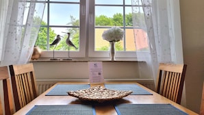 Un comedor con una mesa dispuesta para dos personas, un florero con flores blancas y dos esculturas de pájaros en el alféizar de la ventana.