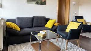 Un salon moderne avec un canapé gris, des coussins jaunes, une table basse en verre et une porte en bois.