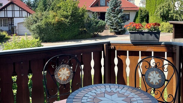 Une terrasse avec une table en mosaïque et deux chaises, entourée d’une clôture en bois et offrant une vue sur des maisons aux toits rouges.