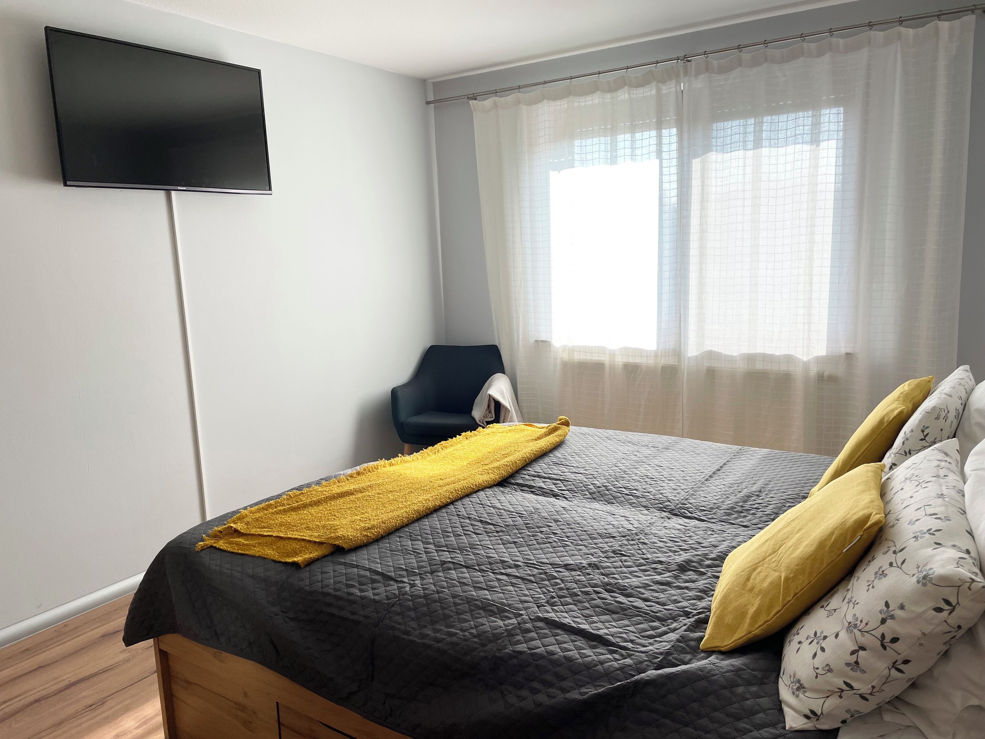 A bedroom with a bed, a chair, a TV, and a window with curtains.
