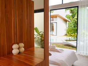 A modern hotel room with a wooden desk, a bed with white linens, and a view of a garden and building outside. - Villa Palmira - 2 bedroom Bali style Pool Villa (Bo Phut)