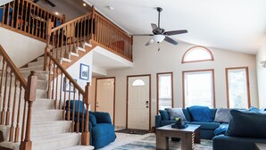 A spacious living room with a wooden staircase, a ceiling fan, and a large window providing natural light.