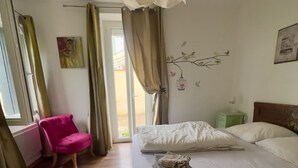 A bedroom with a bed, a chair, a window with curtains, and a wall decoration. - Apartment n°3 - T2 bedroom & separate bathroom (Lamalou-les-Bains)