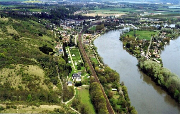 Luftaufnahme einer Stadt an einem Fluss mit einem markanten Haus, einer Bahnlinie und umgebender Natur.