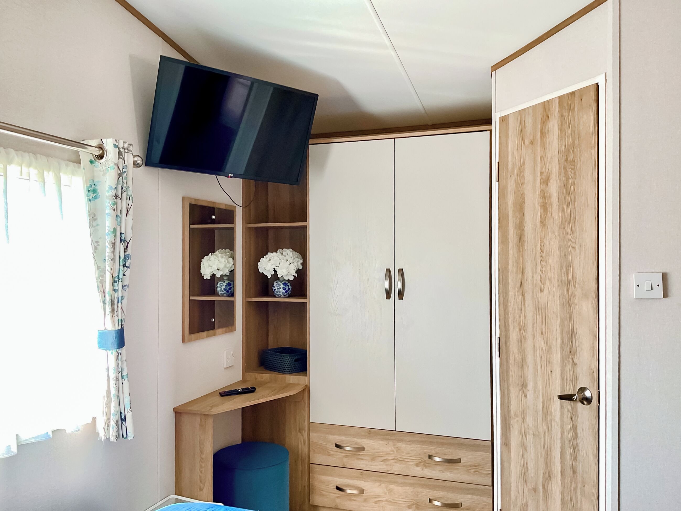 Un dormitorio con televisor montado en la pared, armario de madera y una pequeña estantería con flores.