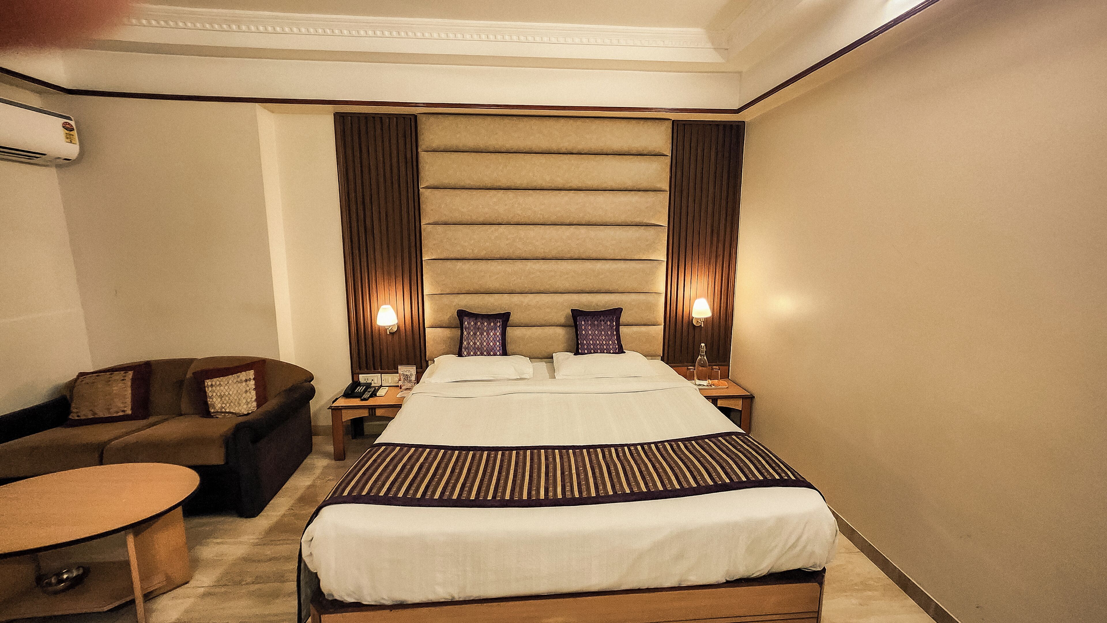 A hotel room with a large bed, a sofa, a bedside table with a lamp, and an air conditioning unit.