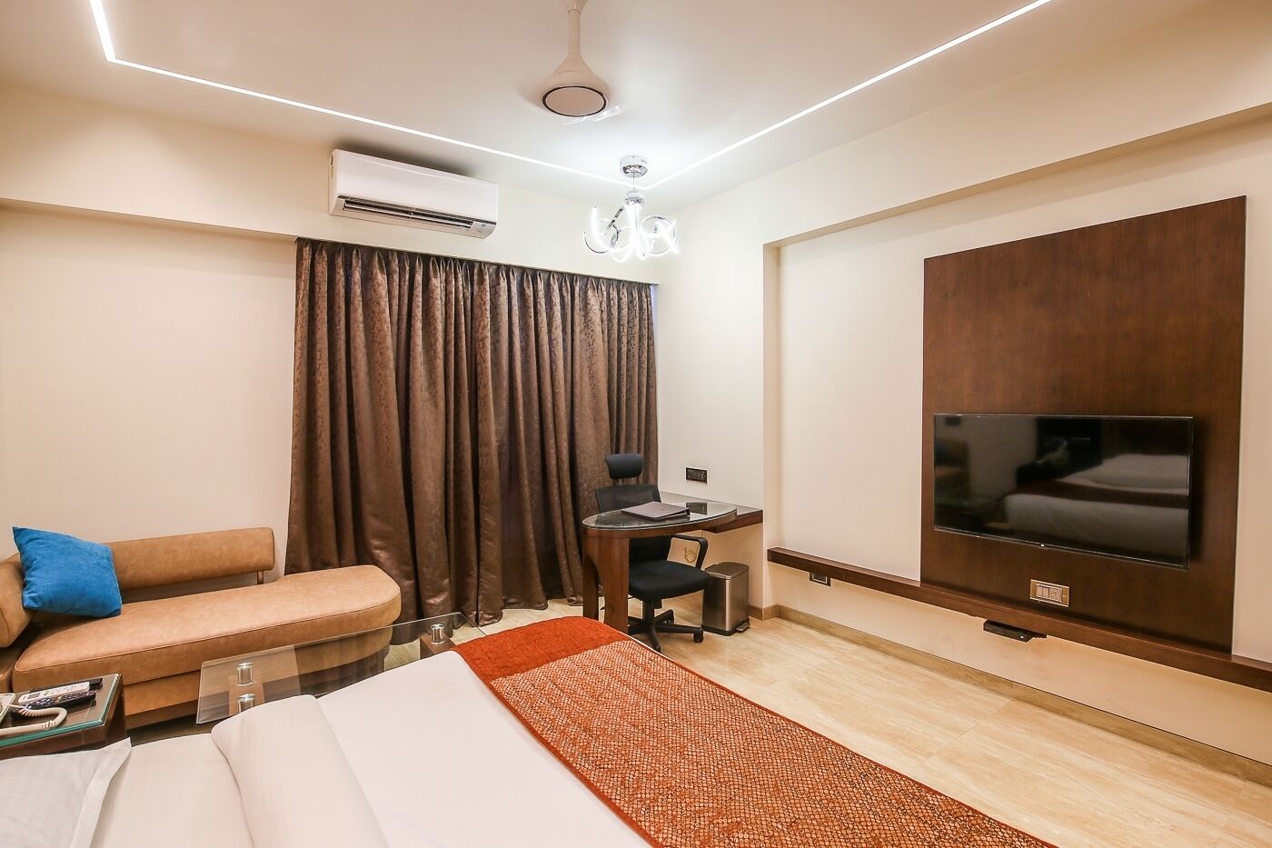 A modern hotel room with a bed, a desk, a chair, a TV, and an air conditioning unit.