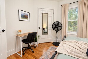 Une chambre à coucher avec un lit, un bureau et une chaise, un ventilateur sur pied, une porte et une fenêtre avec des rideaux.