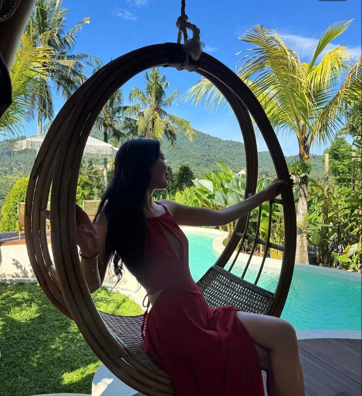 A person sitting in a wicker swing chair overlooking a pool with palm trees and a mountainous backdrop.