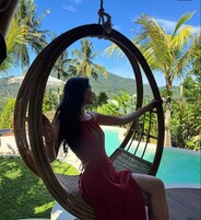 A person sitting in a wicker swing chair overlooking a pool with palm trees and a mountainous backdrop.