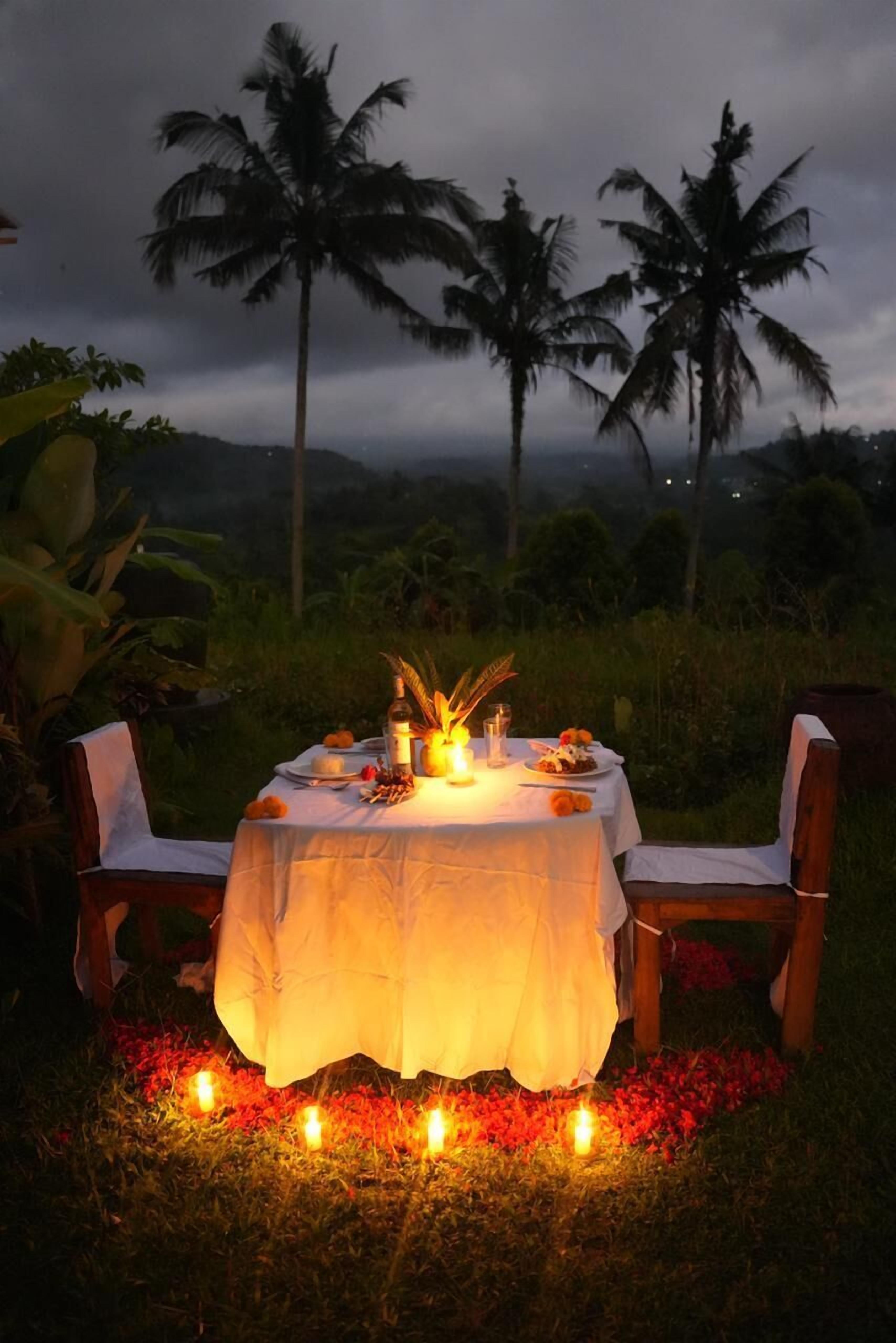 An outdoor dining setup with a table and chairs, candles, and a tropical backdrop.