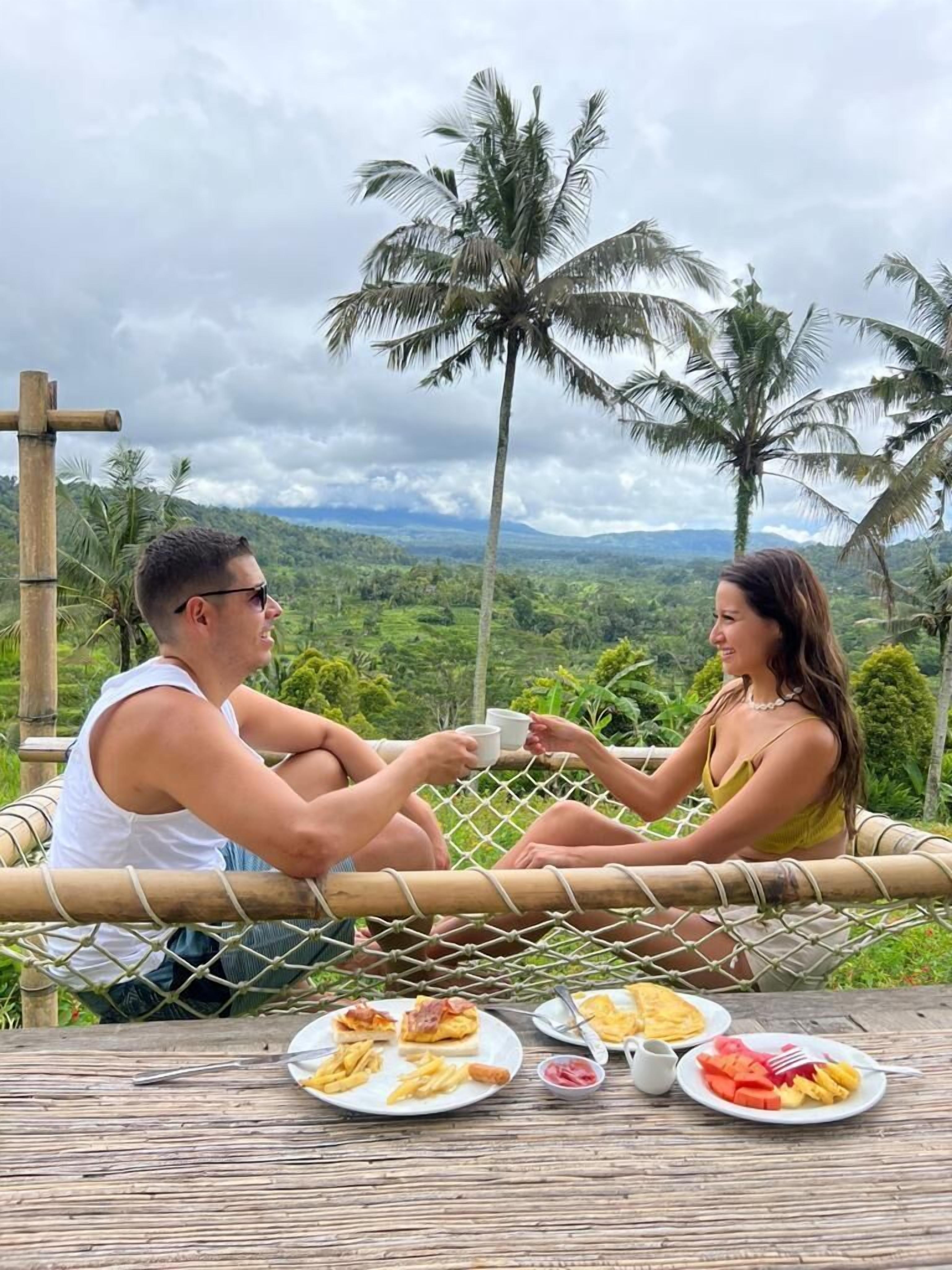 Two people sitting on a hammock with a table of food in front of them, overlooking a lush landscape with palm trees and a cloudy sky.