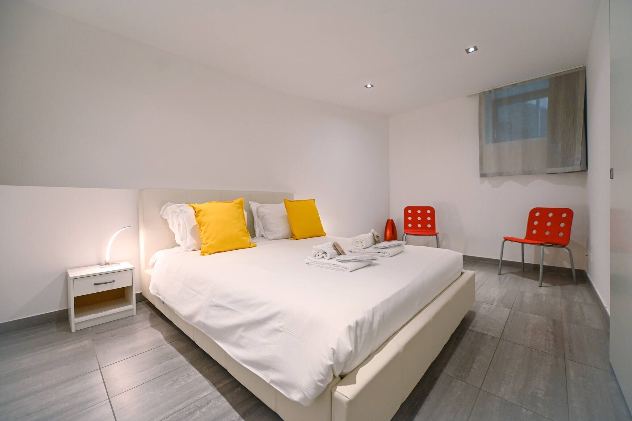 A modern bedroom with a large bed, white bedding, and bright yellow pillows. There is a nightstand with a lamp, a red chair, and a window with a view.
