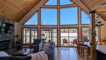A spacious living room with a stone fireplace, large windows, and a wooden ceiling.