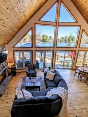 Ein gemütliches Wohnzimmer mit Kamin aus Stein, einer schwarzen Couch und einem Couchtisch. Große Fenster bieten Blick auf eine Terrasse und Gartenmöbel.