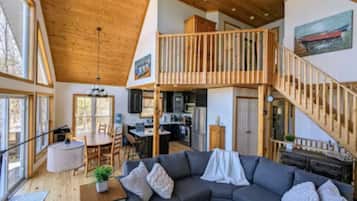 A spacious living area with a wooden ceiling, a large gray sofa, a dining table, and a kitchen in the background.