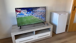 En tv-apparat står på ett TV-bord i trä med två lådor, bredvid ett vitt kylskåp, och golvet är också av trä.
