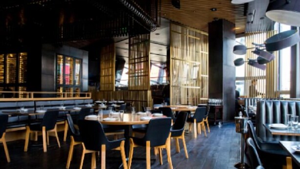 Ein modernes Restaurant mit Holztischen und schwarzen Stühlen, großen Fenstern und einer hohen Decke mit freiliegenden Balken.