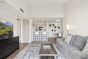 Una sala moderna con un sofá gris, una mesita de centro y un televisor sobre un mueble negro. Se ve una cocina con gabinetes blancos y un salpicadero con diseño de estrellas al fondo.