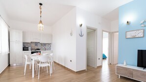 A modern kitchen with white cabinets and a tiled backsplash, a dining area with white chairs, and a living space with a TV and a blue accent wall. - ApartHotel Capo Rizzuto - Sea View Apartments 40mt from the Reserved Beach (Capo Rizzuto)