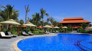 A swimming pool with lounge chairs and umbrellas, surrounded by palm trees and a building with a red roof. - Phi Yen Eco Resort (Can Tho)