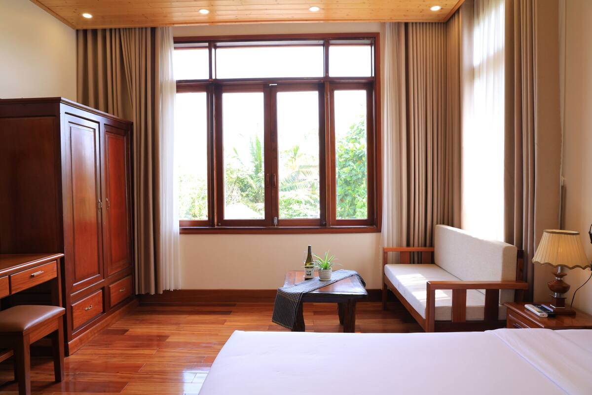 Habitación de hotel con cama, escritorio, silla, una mesita con una botella y planta, y una ventana con cortinas.