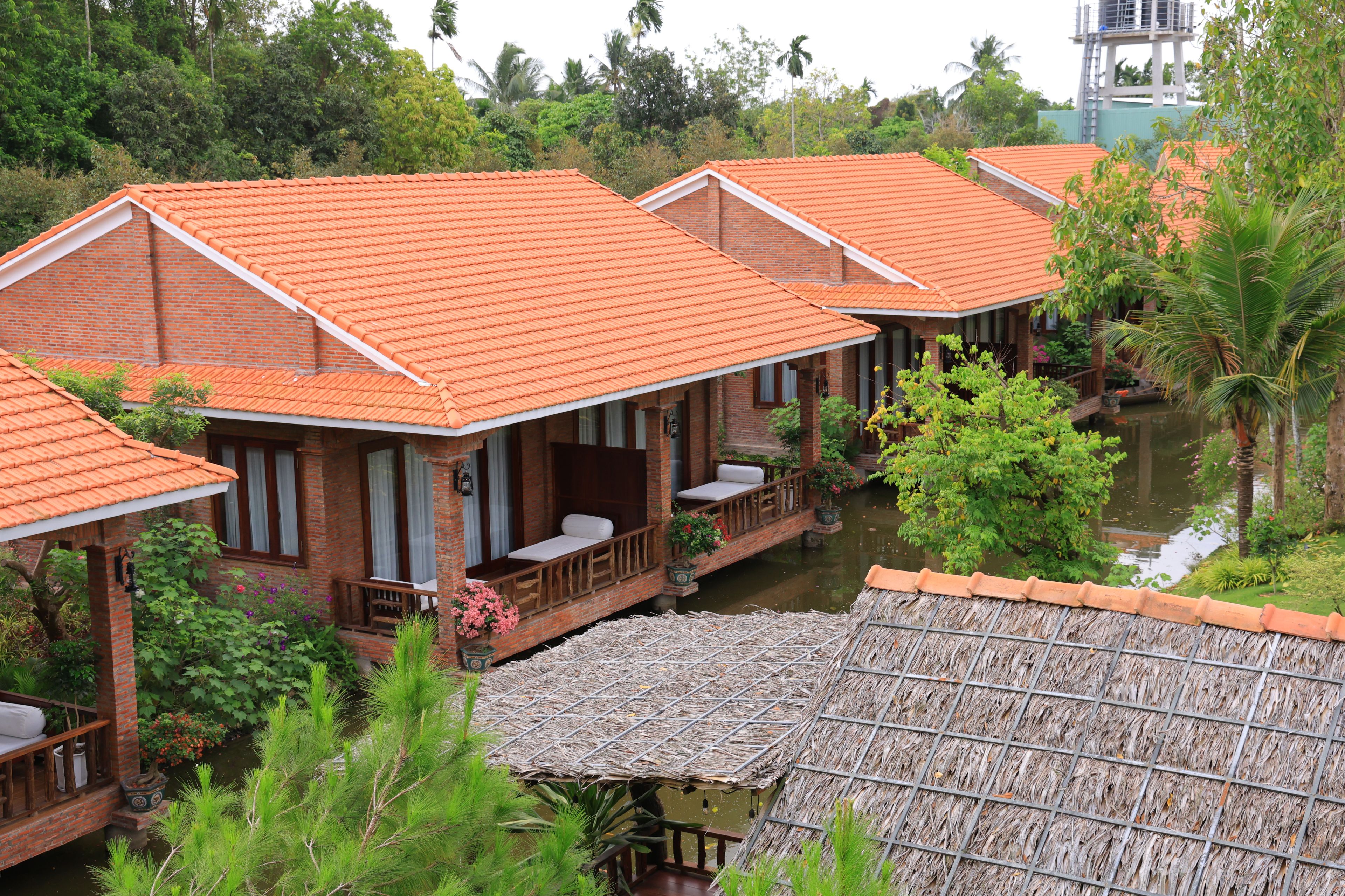 A row of brick buildings with orange tiled roofs, a thatched-roof structure, and a water body in the foreground.