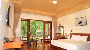 A hotel room with a wooden ceiling, a large bed, a desk with a telephone, a table with chairs, a nightstand with a lamp, and a large window with a view of greenery. - Phi Yen Eco Resort (Can Tho)