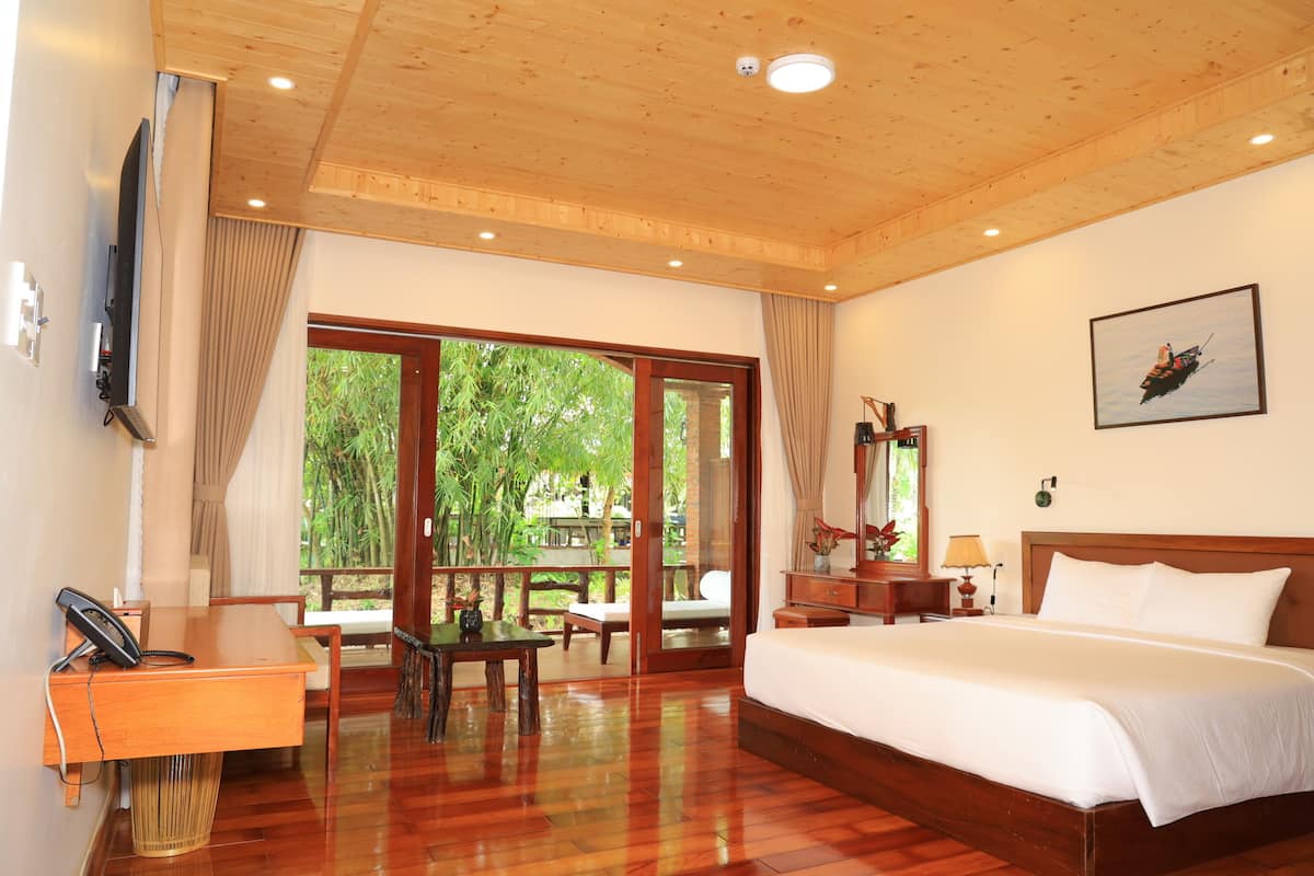Habitación de hotel con techo de madera, una cama grande, un escritorio con teléfono, una mesa con sillas, una mesita de noche con lámpara y un ventanal con vistas a zonas verdes.