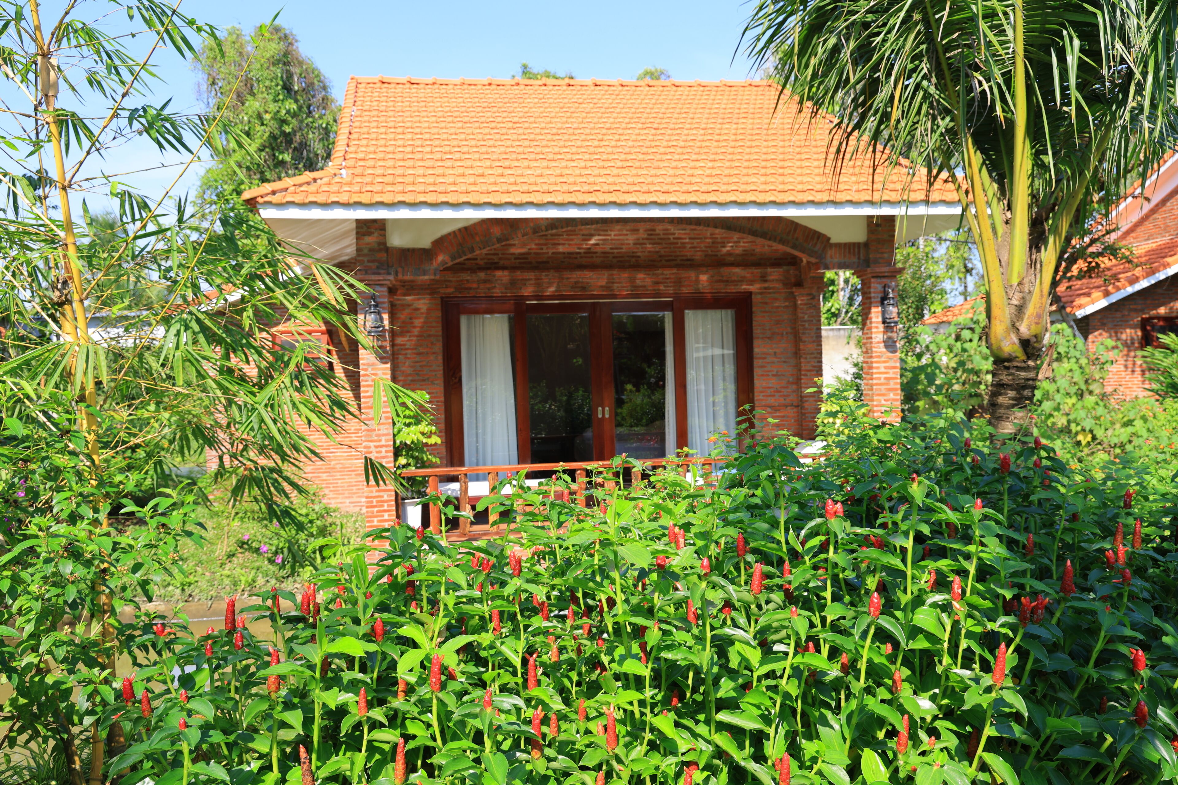 Une maison en briques avec un toit en tuiles, entourée d’une végétation luxuriante et de fleurs rouges.