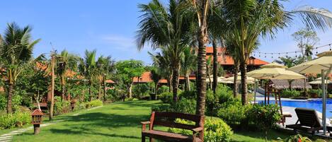 A lush garden with a wooden bench, palm trees, a swimming pool, and thatched-roof buildings.