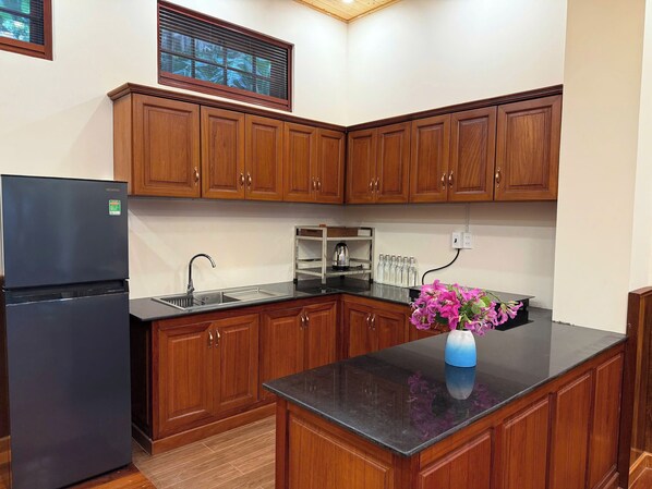 A kitchen with wooden cabinets, a black refrigerator, a stainless steel sink, and a vase with flowers on the counter. - Phi Yen Eco Resort (Can Tho)