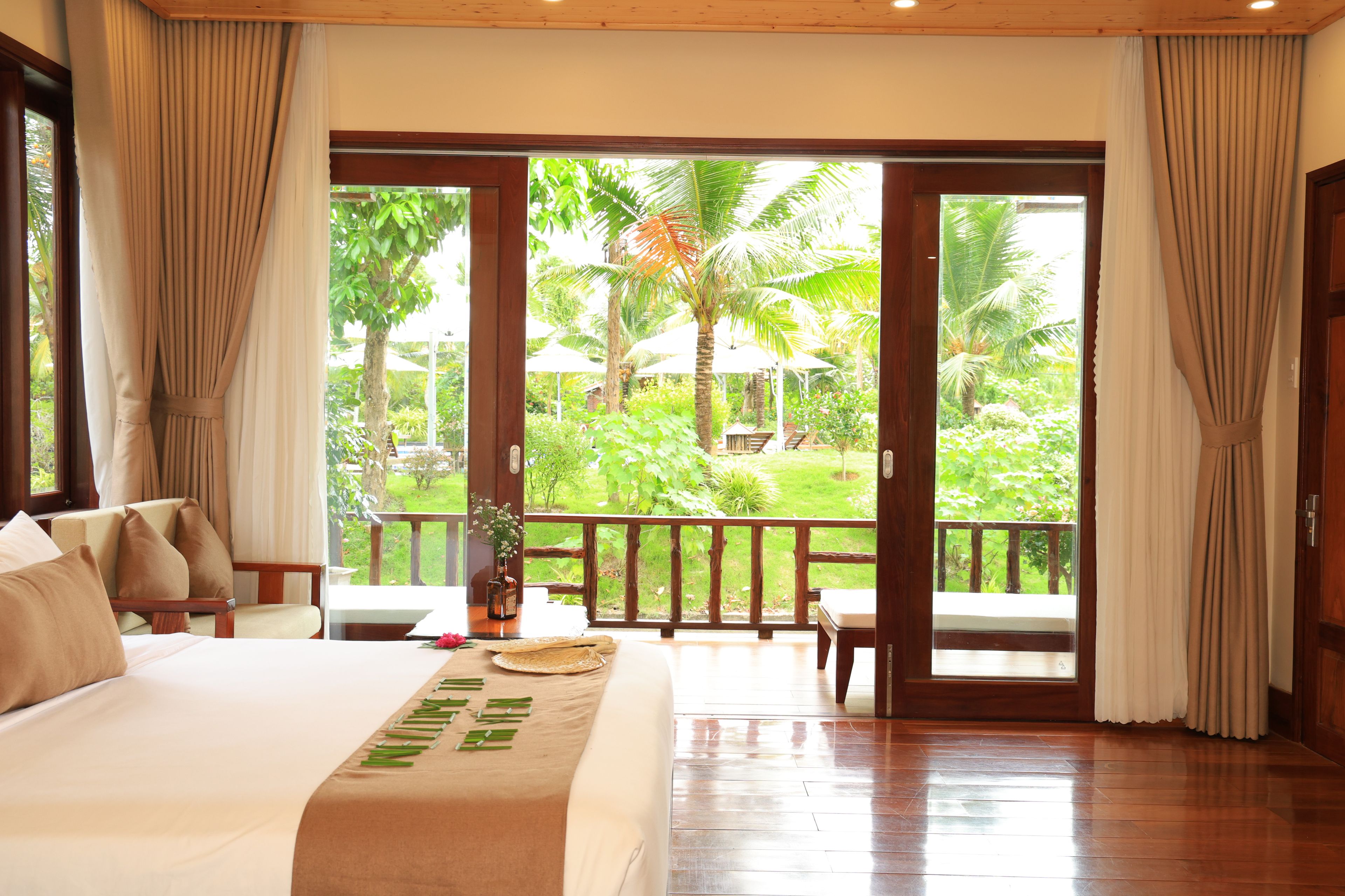 A hotel room with a large bed, a view of a tropical garden, and a balcony with a wooden railing.