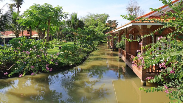 A canal with a wooden bridge, lush greenery, and a building with a tiled roof. - Phi Yen Eco Resort (Can Tho)
