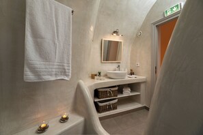 A bathroom with a white sink, mirror, and towel hanging on the wall. There are baskets and shelves below the sink. - Messavoli residences (Santorini)