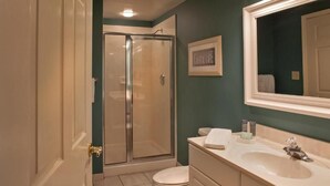 A bathroom with a glass shower enclosure, a toilet, a sink with a mirror, and a framed picture on the wall.