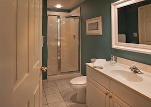 A bathroom with a glass shower enclosure, a toilet, a sink with a mirror, and a framed picture on the wall.