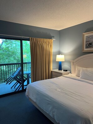 Habitación de hotel con cama, mesita de noche con lámpara, ventana con cortinas y vista a zonas verdes.