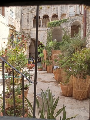 Une cour agrémentée de plantes en pot, dotée d’un espace extérieur aménagé pour s’asseoir, et un bâtiment en pierre orné d’arcades.