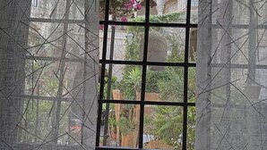 A window with a metal grid and sheer curtains, offering a view of a building and greenery outside. - Very nice little apartment in the heart of the beautiful island of Ortigia (Siracusa)