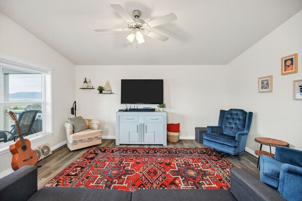 A living room with a ceiling fan, a flat-screen TV, a blue armchair, a red patterned rug, and a guitar.
