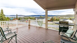 A covered outdoor deck with a grill, two chairs, and a view of a residential area with houses and mountains in the background.