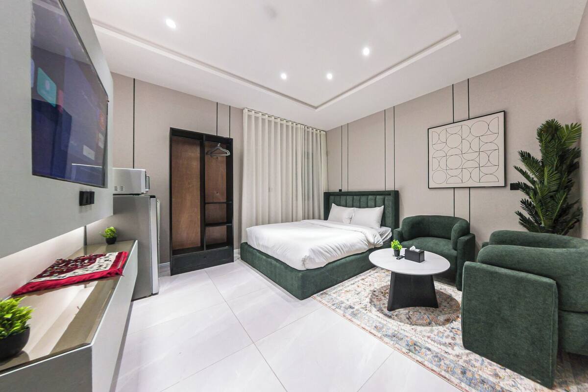 Moderni hotellihuone, jossa on sänky, vihreät nojatuolit, pieni pyöreä pöytä ja suuri ruukussa oleva kasvi.