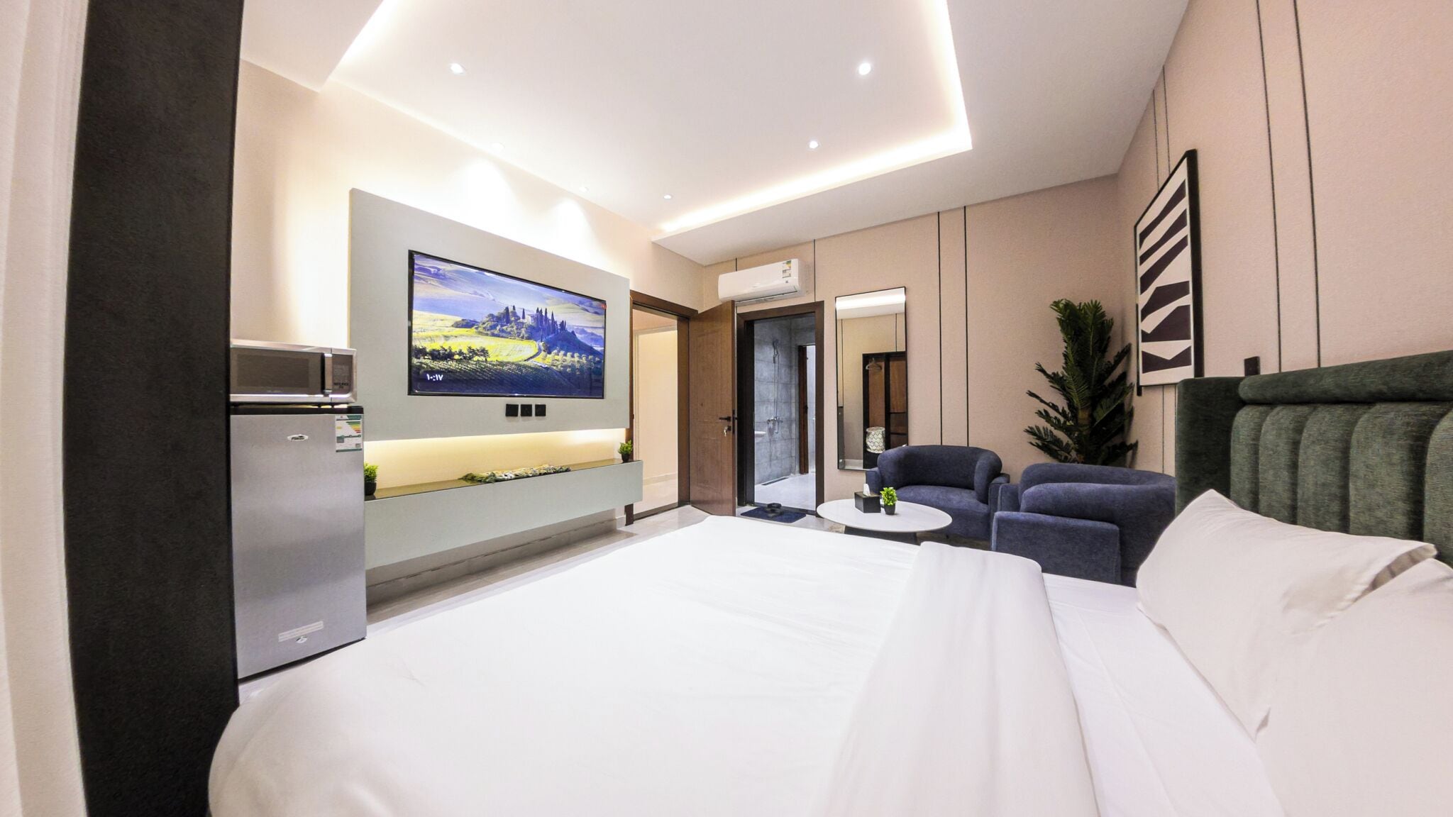A modern hotel room with a large bed, a flat-screen TV mounted on the wall, a small seating area with two chairs, and a built-in air conditioning unit.
