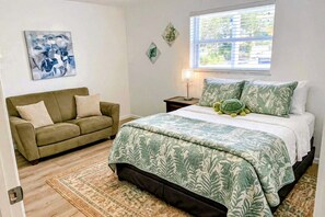 A bedroom with a bed, a sofa, a nightstand, a lamp, and a window with blinds.