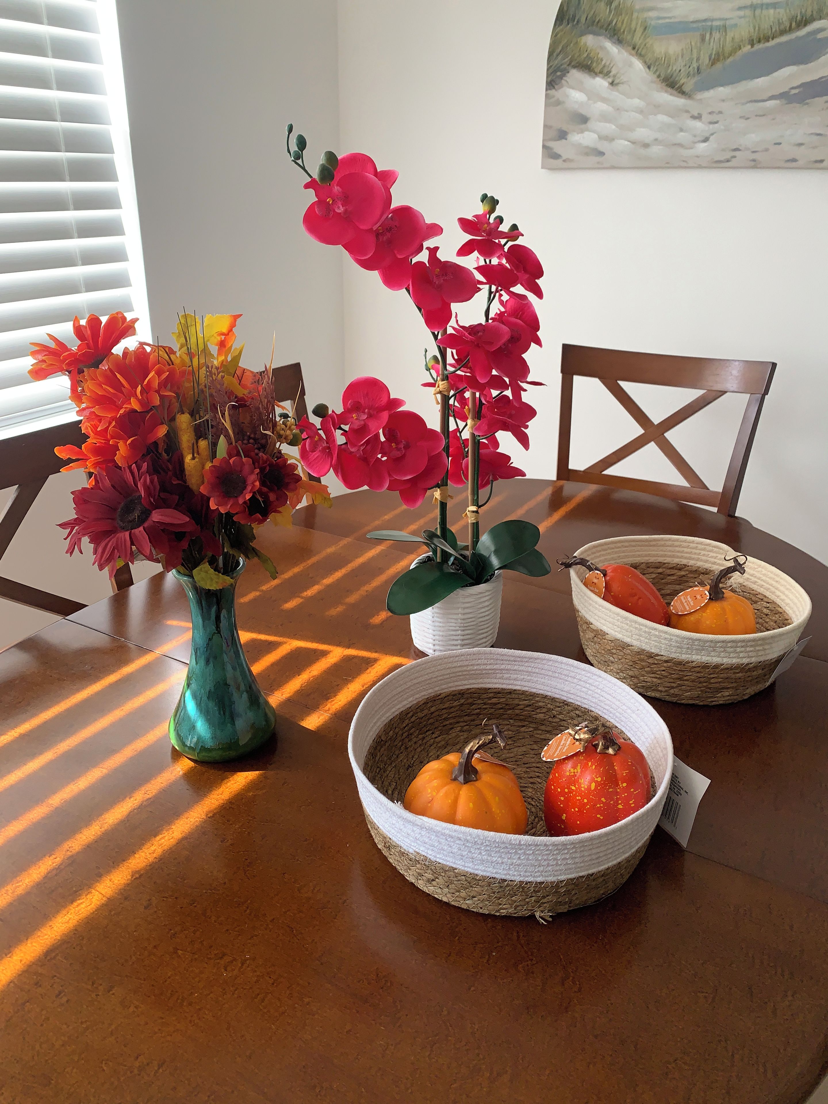 A dining table with a vase of flowers, a potted plant, and woven baskets containing pumpkins and a tomato.